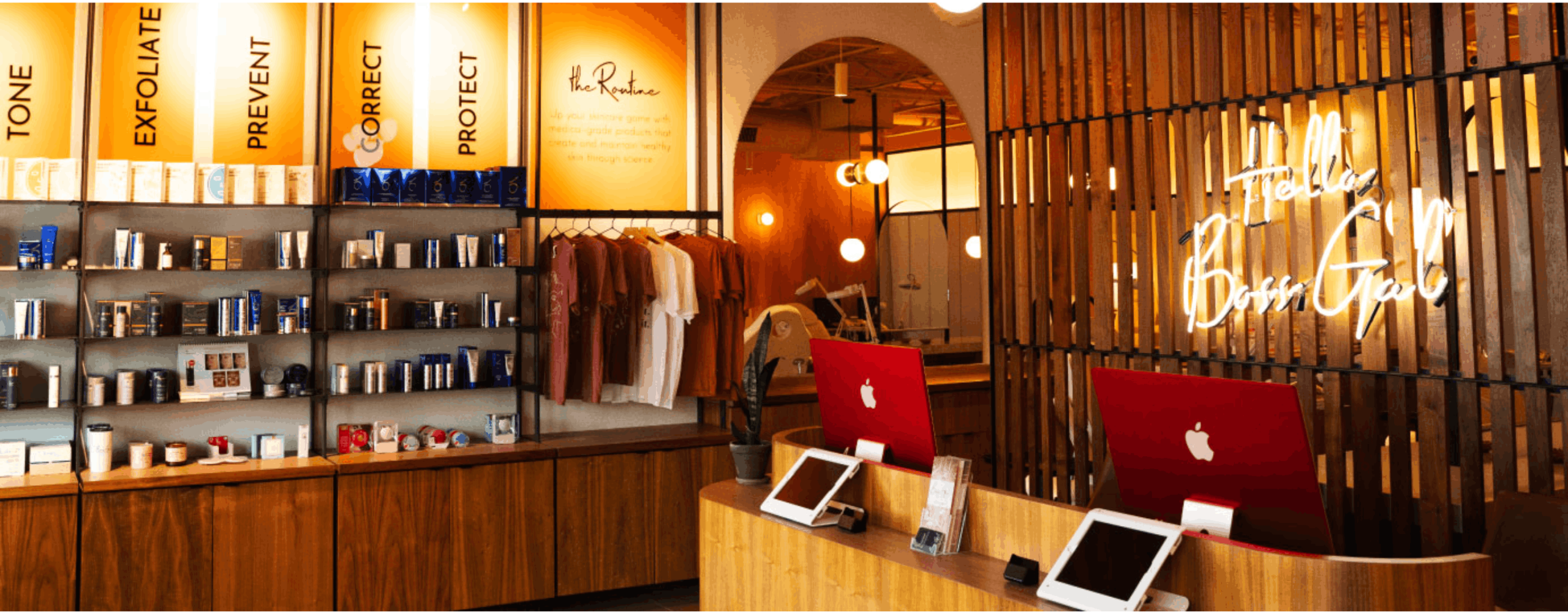 A chic medical spa interior features wooden cabinetry displaying skincare products underneath lighted panels, next to a rack of t-shirts and a wooden slat accent wall behind the reception desk.