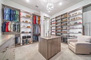 A spacious, luxury custom walk-in closet featuring light wood cabinetry, extensive built-in shoe racks, a central island, a chandelier, and a plush armchair in the corner.