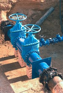 Large blue industrial water valves and steel piping are installed in a trench supported by brick stacks during an outdoor commercial plumbing construction project.
